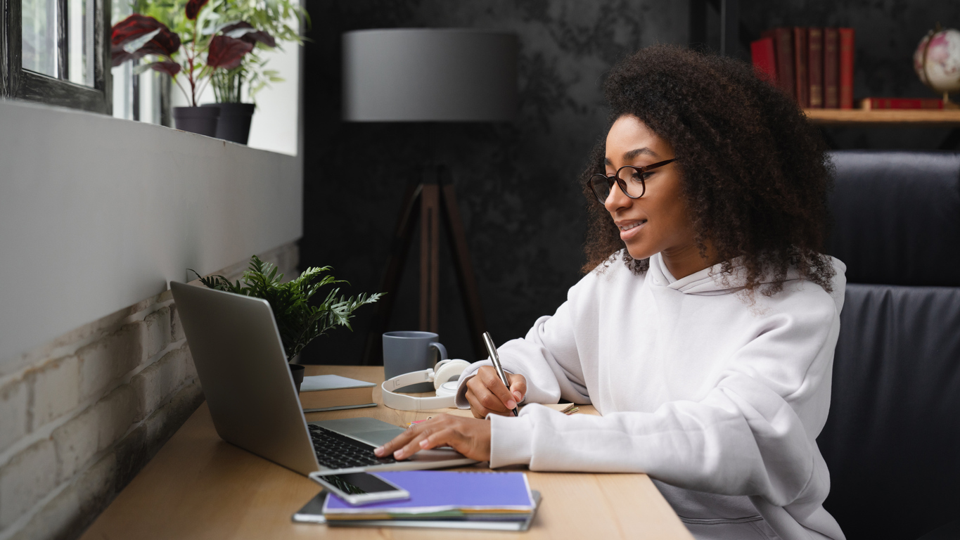 Graduate student working on laptop and taking notes at desk in their apartment