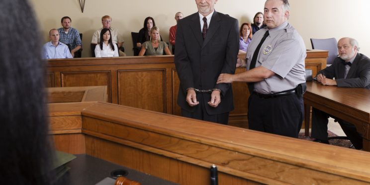 Handcuffed defendant stands with bailiff in courtroom