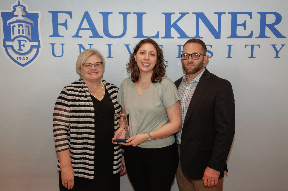 l-r Cathy Davis, Mary Beth and John Simmons pose in front of a Faulkner backdrop. Mary Beth holds a Distinguished Alumna award.