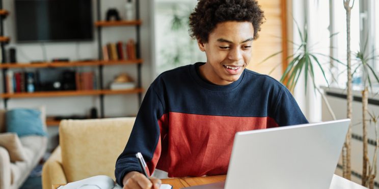 Student smiles while engaging with laptop computer screen and taking notes by hand at home