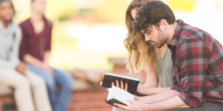 Two college students reading Bible together on park bench outside